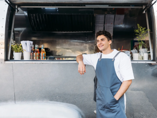 Young business owner in an apron. Salesman leaning on a food truck.