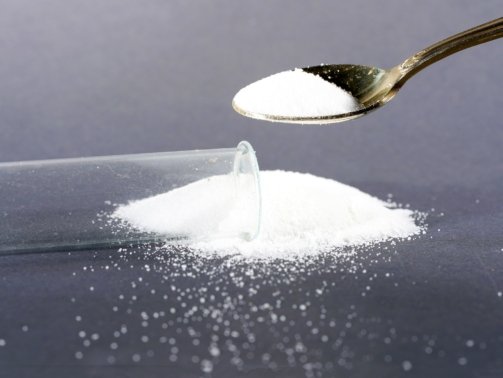 Metal spoon stuffing white powder into a capsule in a lab with scattered grains