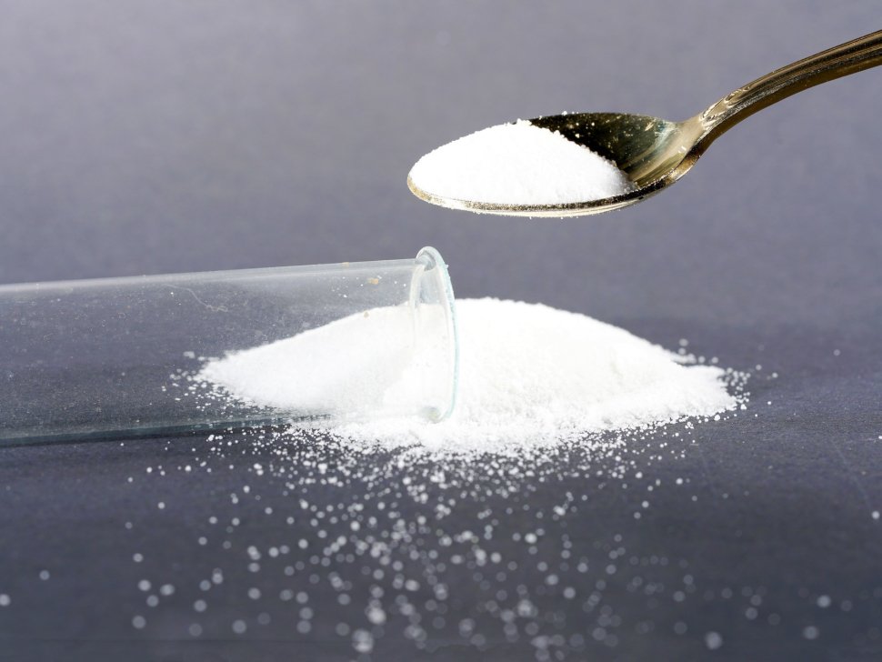 Metal spoon stuffing white powder into a capsule in a lab with scattered grains