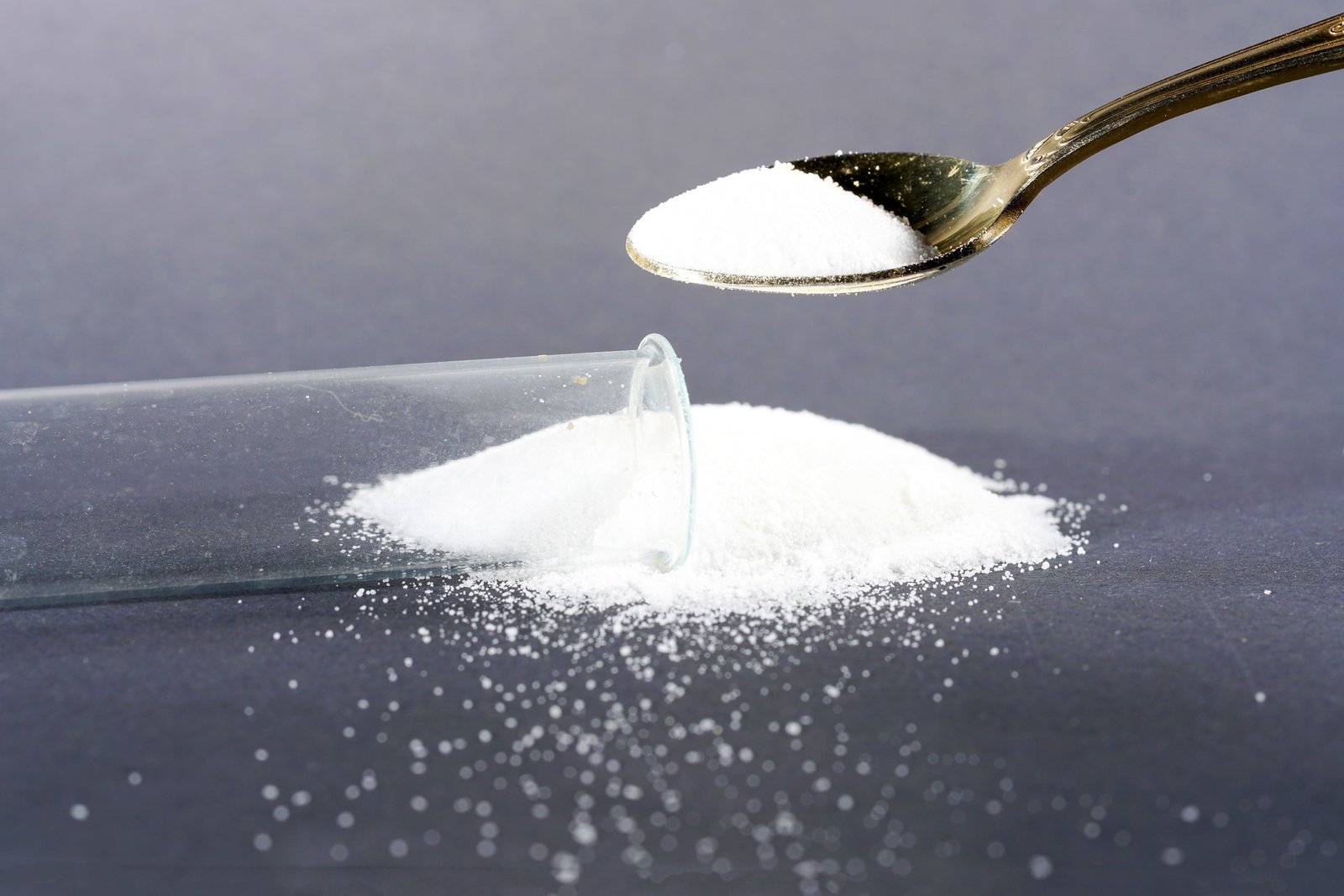 Metal spoon stuffing white powder into a capsule in a lab with scattered grains